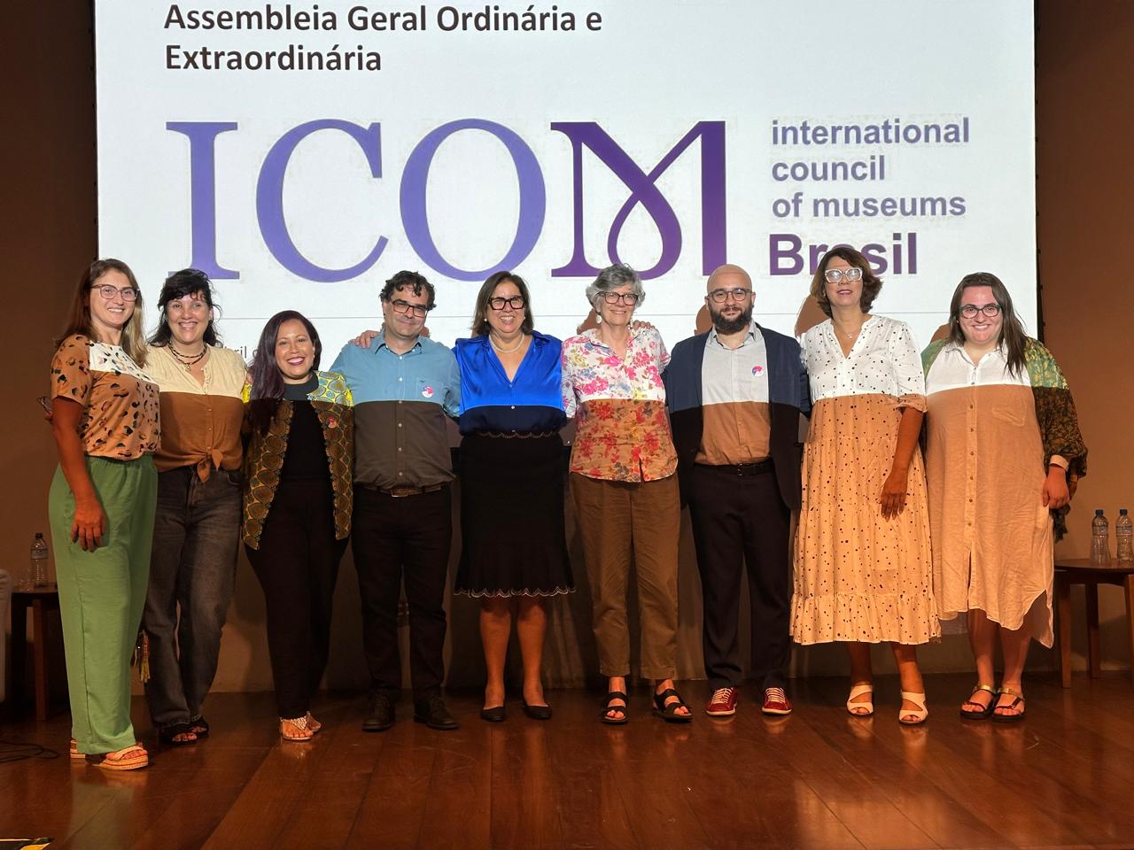 oito pessoas abraçadas em frente à uma projeção no qual está escrito Assembleia Geral Ordinária e Extraordinária ICOM Internation Council of Museums Brasil