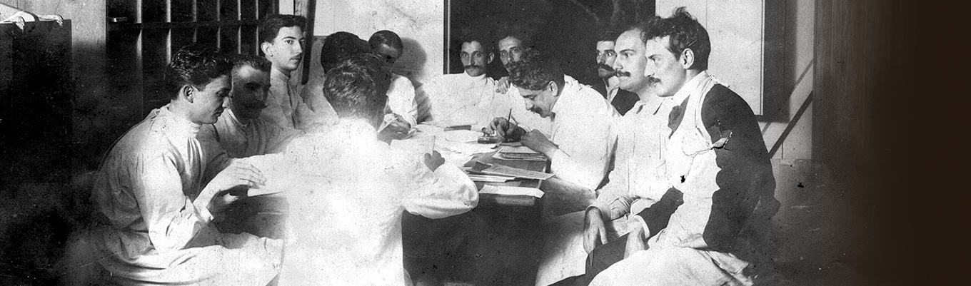 Grupo de cientistas reunidos em uma mesa com papéis sobre ela. Oswaldo Cruz faz anotações.
