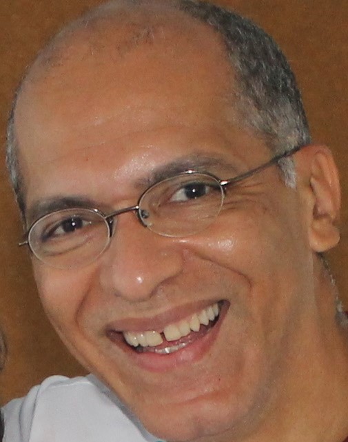 Homem branco, calvo, grisalho, de óculos, em primeiro plano, sorrindo.