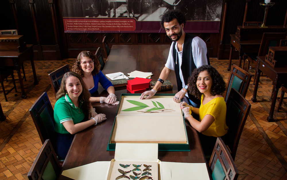 Estudantes - três mulheres e um homem ' ao redor de uma mesa, sorrindo, com livros abertos mostrando ilustrações de plantas.