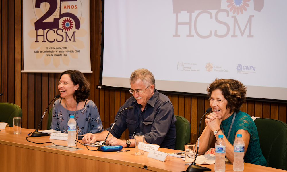 Três palestrantes, sendo uma mulher, um homem e outra mulher, compõem a mesa. Eles sorriem. Ao fundo, um cartaz diz: HCSM 25 anos.