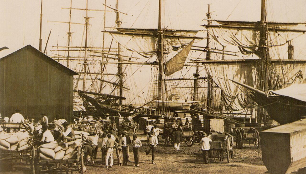 Foto antiga do Porto do Santos. Vê-se um galpão, com muitos barcos à vela ao fundo e muitas pessoas em primeiro plano.