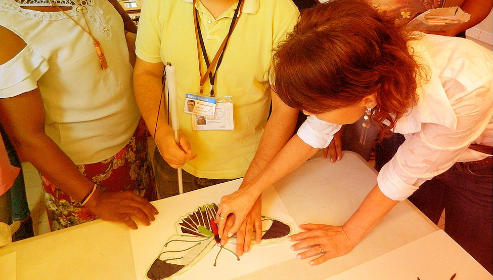 Homem cego toca em uma instalação tátil representando uma borboleta sobre uma mesa, com a ajuda de uma mulher
