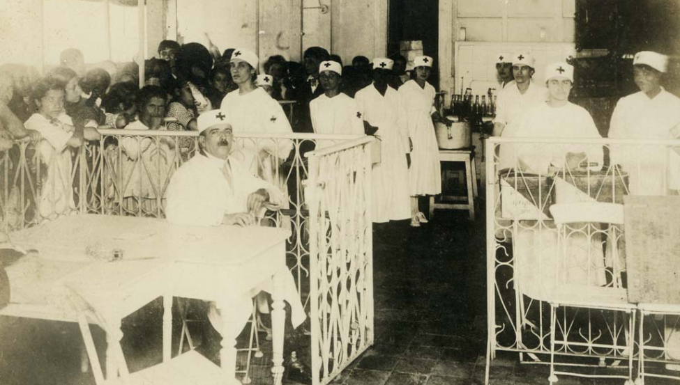 Diversos leitos de hospital com enfermeiros ou médicos com uniforme e chapéu branco com uma cruz, ao fundo pacientes, a maioria crianças