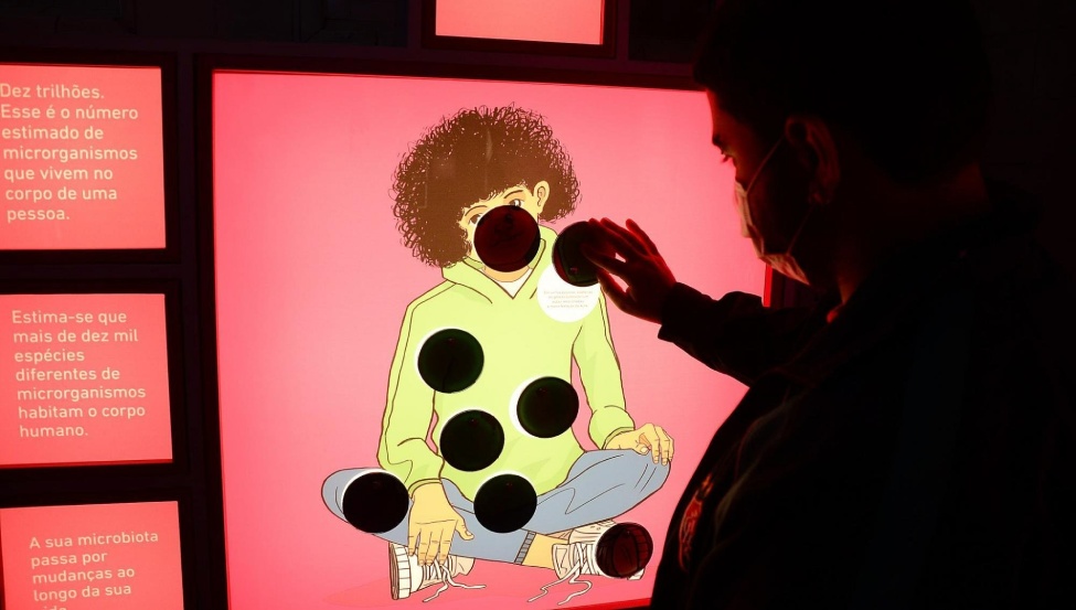 Mulher interage com módulo que traz um painel retroiluminado, no qual se vê a ilustração de uma mulher, tocando-o.