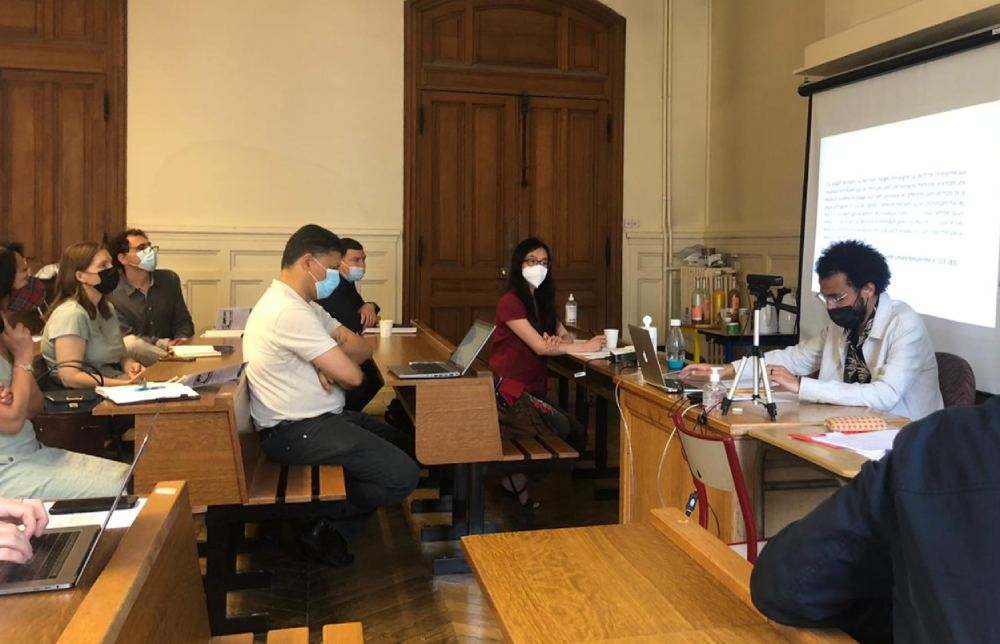José Roberto está sentado à mesa à frente da turma em uma sala de aula. Ele fala enquanto é observado por homens e mulheres com máscaras sanitárias.
