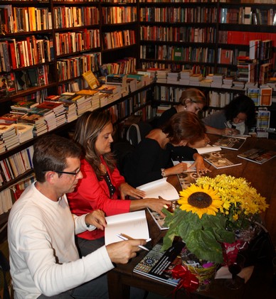 cercados por estantes de livros, autores sentados em uma mesa autografam o livro