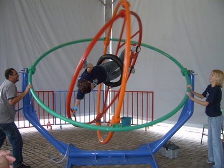 Criança presa no centro do aparelho da Nasa, que simula falta de gravidade, manuseado por duas pessoas que se encontram ao lado. 