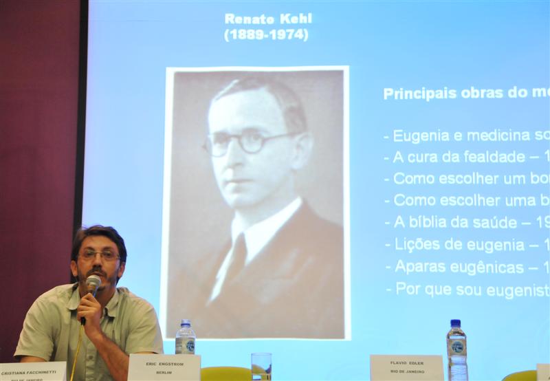 palestrante fala ao microfone. ao fundo, foto em preto e branco se destaca em sua apresentação de power point
