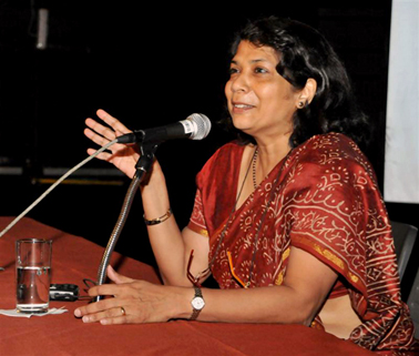 moça morena usando traje típico da India fala ao microfone com as mãos sobre uma mesa
